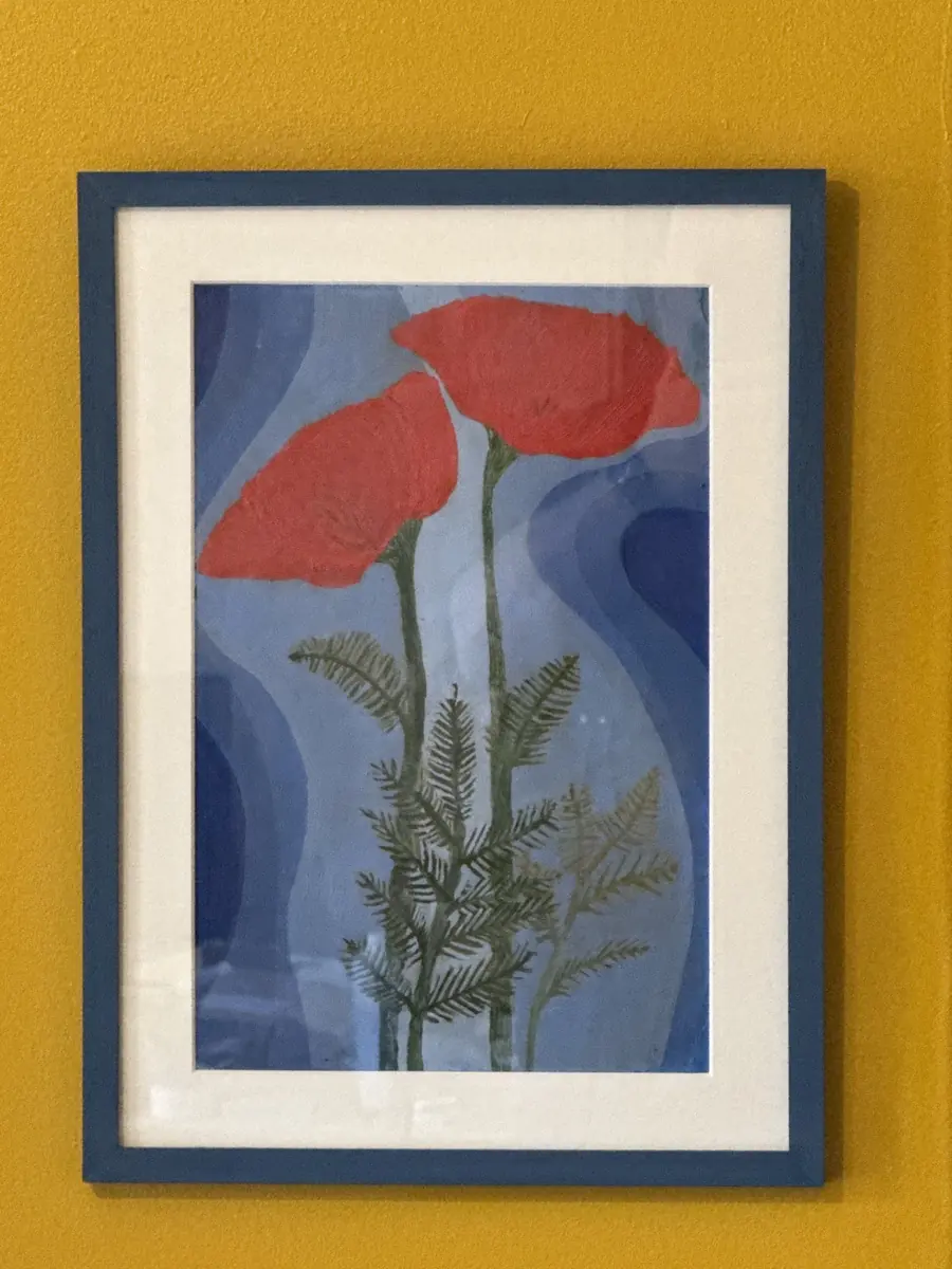 A stylised red poppy print in a dark navy frame with a cream mount, against a bright mustard-yellow background