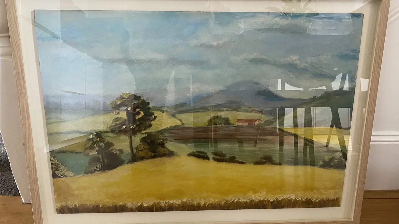 A naive-style landscape painting of a tree, fields and distant hills in a wide pale wood frame, leaning against a white wall